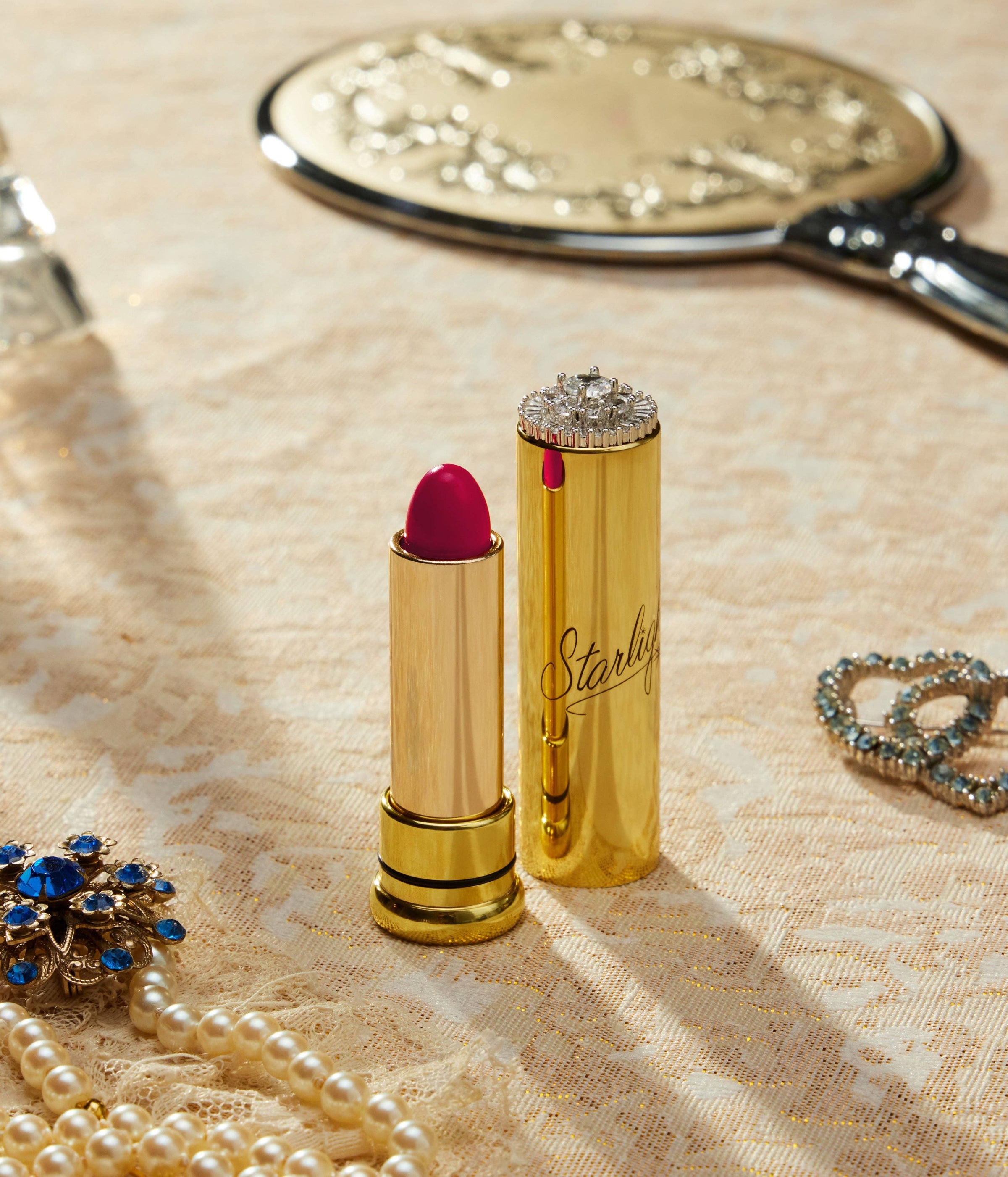 Gold lipstick with red lipstick on a textured surface with jewelry and a mirror.