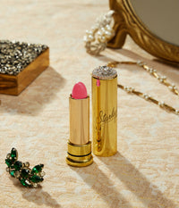 Gold lipstick with pink shade and decorative cap on a textured surface with jewelry.