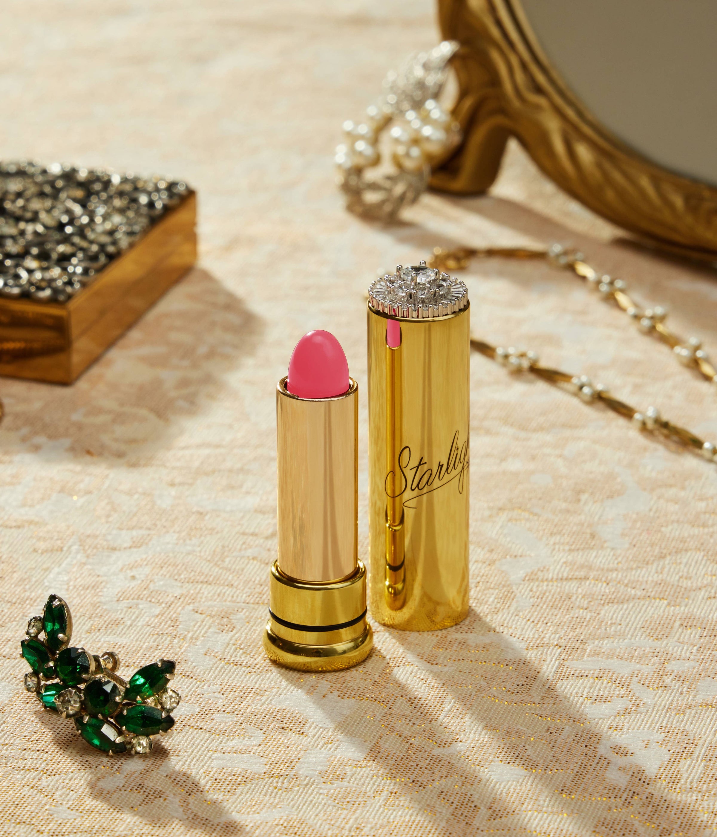 Gold lipstick with pink shade and decorative cap on a textured surface with jewelry.
