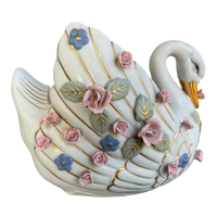 A decorative swan figurine in glossy white porcelain with gold-painted lines, pink roses, blue flowers, and green leaves, isolated on a white background.