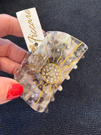 A hand with red nail polish holds a resin hair claw clip featuring a sunburst design with gold lines, a crystal-embellished center, and small pearl-like accents. A white paper tag with the brand name 'Riccare' is attached.