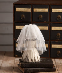 Small figurine of a bride in a white dress with a veil on top of stacked books, with a vintage cabinet in the background.
