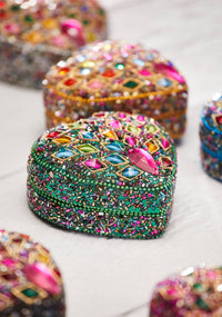 A close-up of a green beaded and rhinestone-embellished heart-shaped decorative piece, with other similar pieces blurred in the background on a light surface.