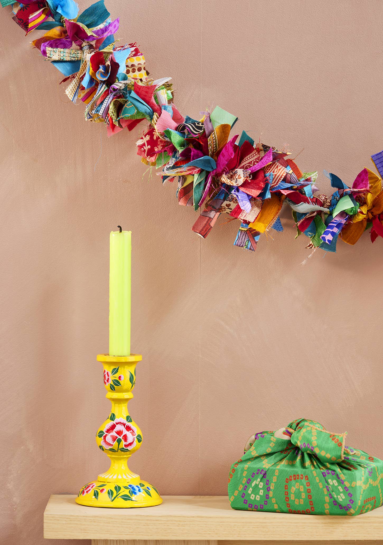 Decorative colorful fabric garland, yellow candlestick with floral design, and green fabric bag on a beige background.