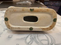 A vintage ceramic soap dish or tray with a central oval cutout, featuring a cream-colored glazed surface with green dots and brown patina around the edges, resting on a patterned tablecloth.