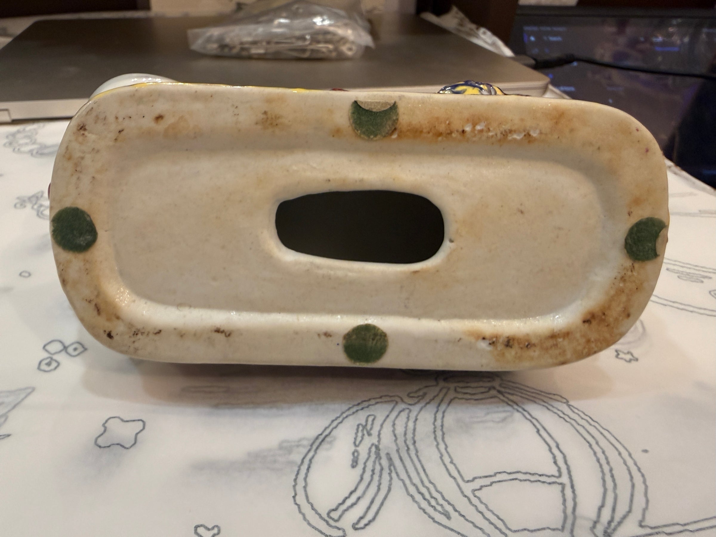 A vintage ceramic soap dish or tray with a central oval cutout, featuring a cream-colored glazed surface with green dots and brown patina around the edges, resting on a patterned tablecloth.