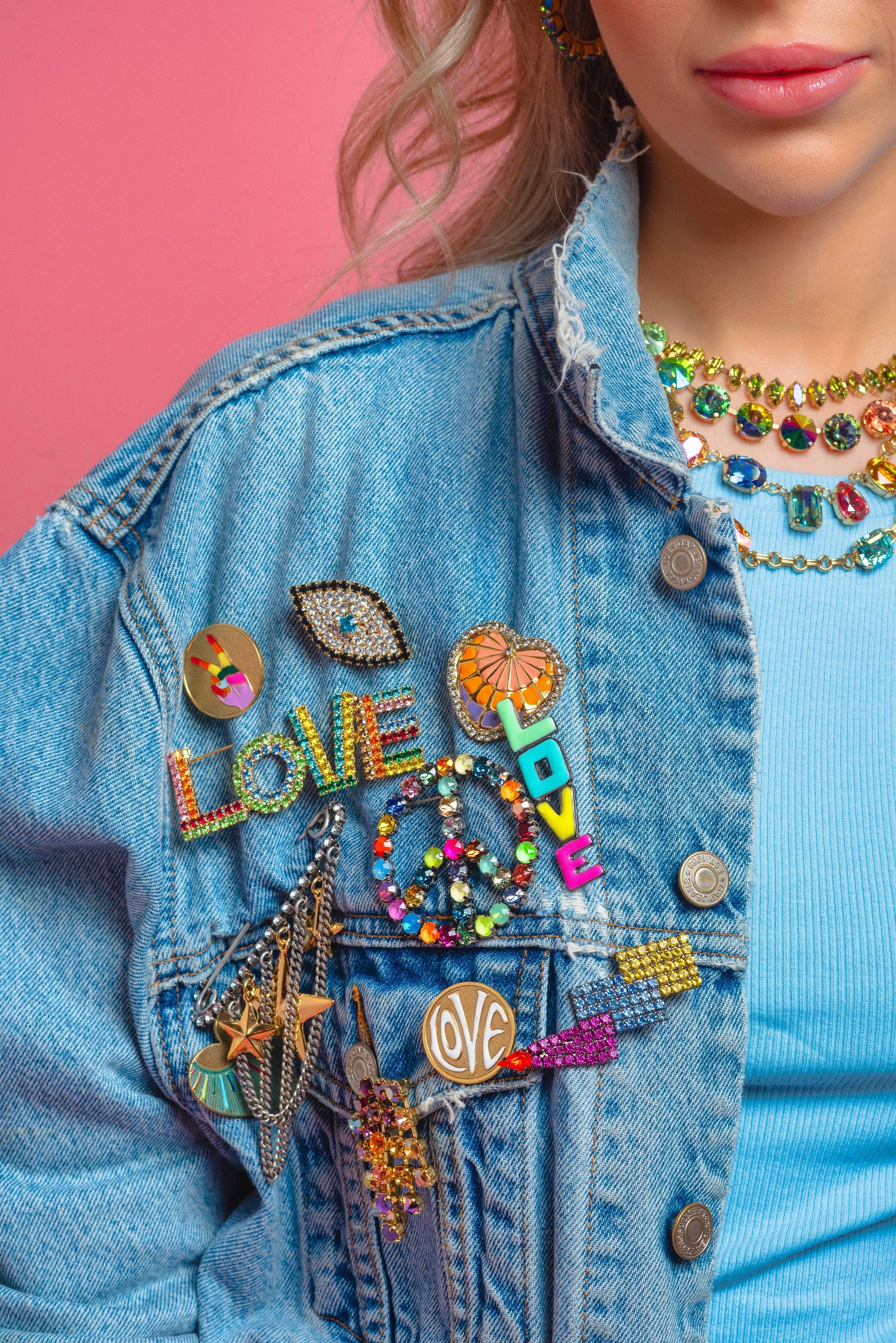 Person wearing a denim jacket with colorful patches and accessories against a pink background