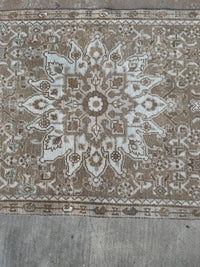A close-up of a vintage-style woven rug with a central medallion and ornate floral patterns in beige, brown, and off-white, laid flat on a gray concrete floor.