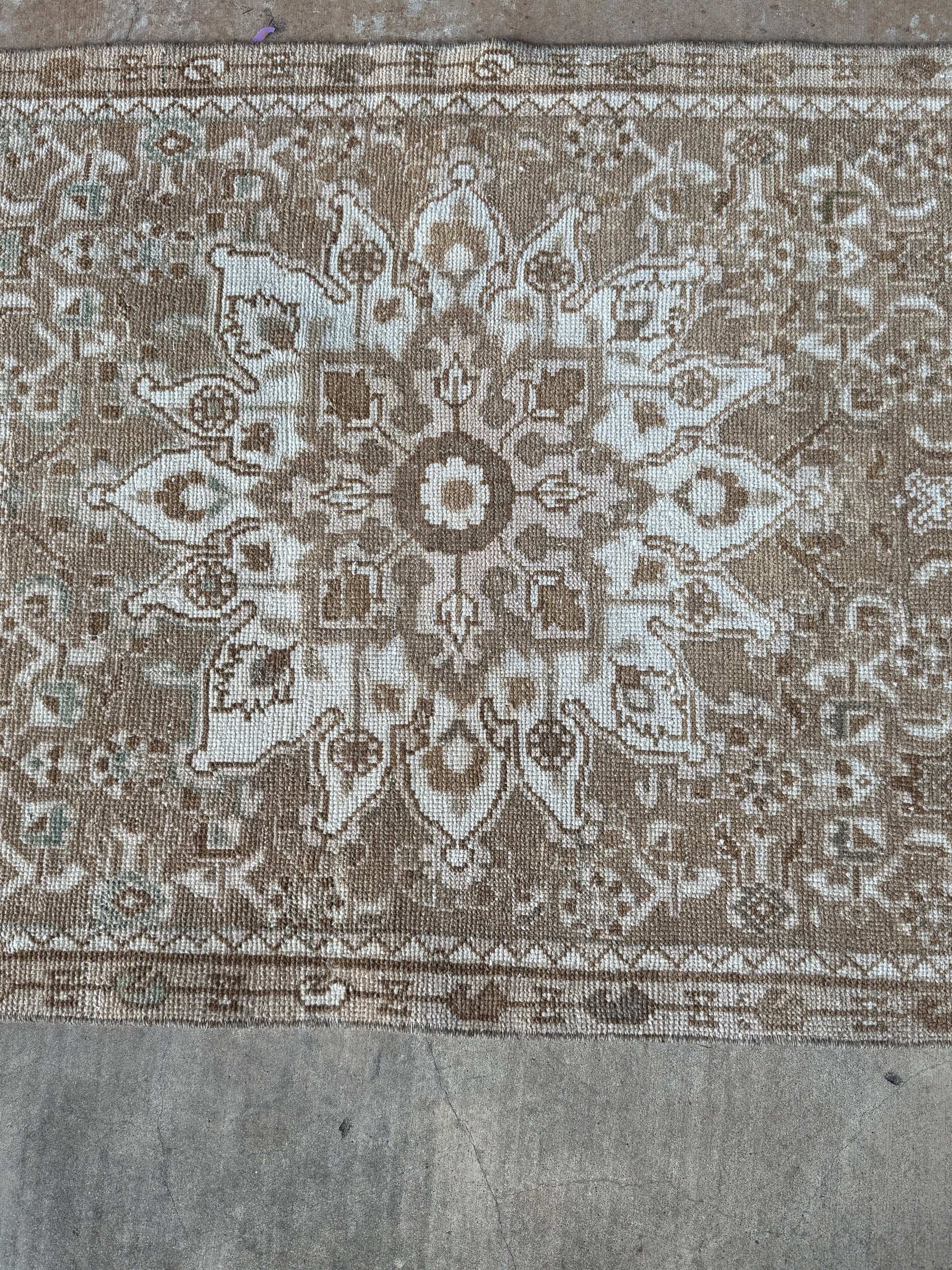 A close-up of a vintage-style woven rug with a central medallion and ornate floral patterns in beige, brown, and off-white, laid flat on a gray concrete floor.