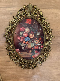 An ornate oval wall art piece with a vintage-style gold frame, featuring a colorful still life floral painting of red, pink, blue, and white flowers in a vase, displayed on a light wood surface.