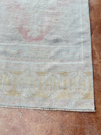 A close-up of a vintage area rug corner showing a faded light gray field with a beige geometric border and a faint pink motif, laid on a brown tiled floor.