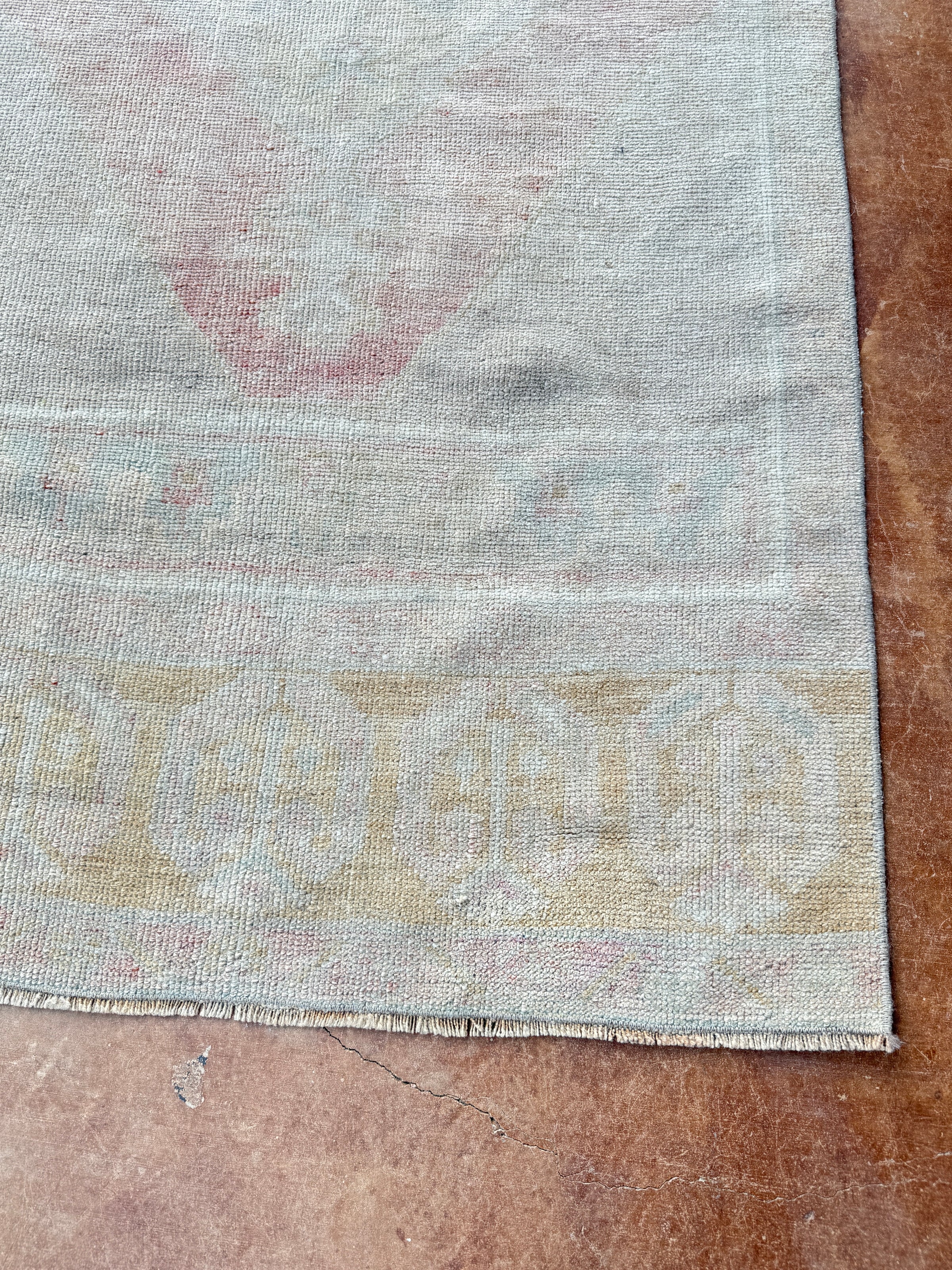 A close-up of a vintage area rug corner showing a faded light gray field with a beige geometric border and a faint pink motif, laid on a brown tiled floor.