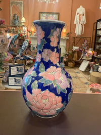 A tall ceramic vase with a cobalt blue base, decorated with large pink and peach flowers and green leaves, displayed on a counter inside a shop.