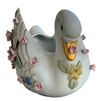 A decorative ceramic swan-shaped planter with a glossy off-white glaze, featuring a gold-painted beak, gold and green leaf details, and pink roses with blue accents on its body and tail.