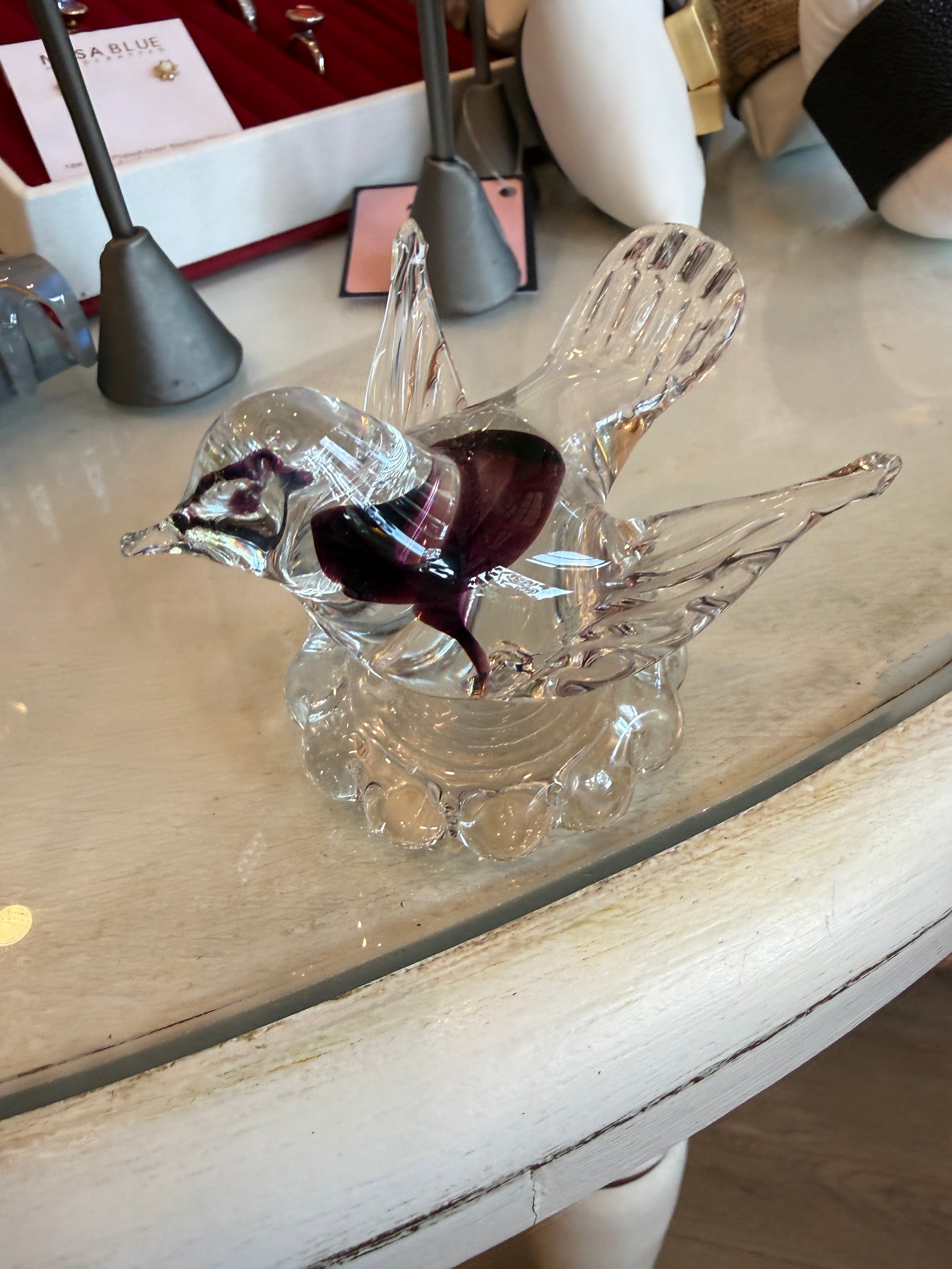 A clear glass bird figurine with a dark purple accent on its chest, displayed on a light-colored wooden table.