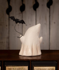A white ceramic ghost figurine with a black glitter bat decoration perched on its shoulder, displayed on a dark wooden cabinet against a rustic wood-paneled background.
