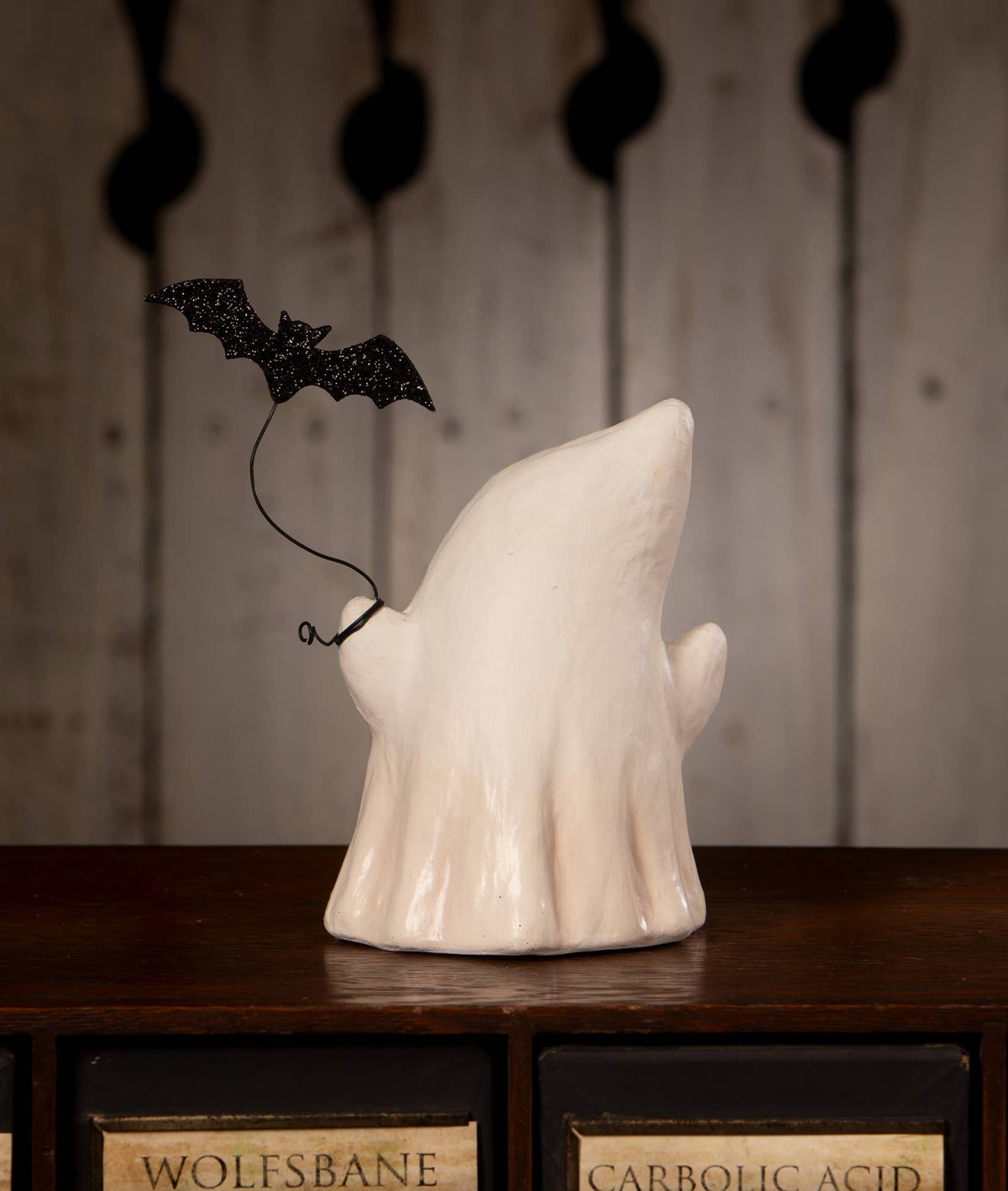 A white ceramic ghost figurine with a black glitter bat decoration perched on its shoulder, displayed on a dark wooden cabinet against a rustic wood-paneled background.