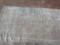 A distressed vintage-style area rug with a faded beige, gray, and muted pink pattern, laid flat on a brown textured floor.
