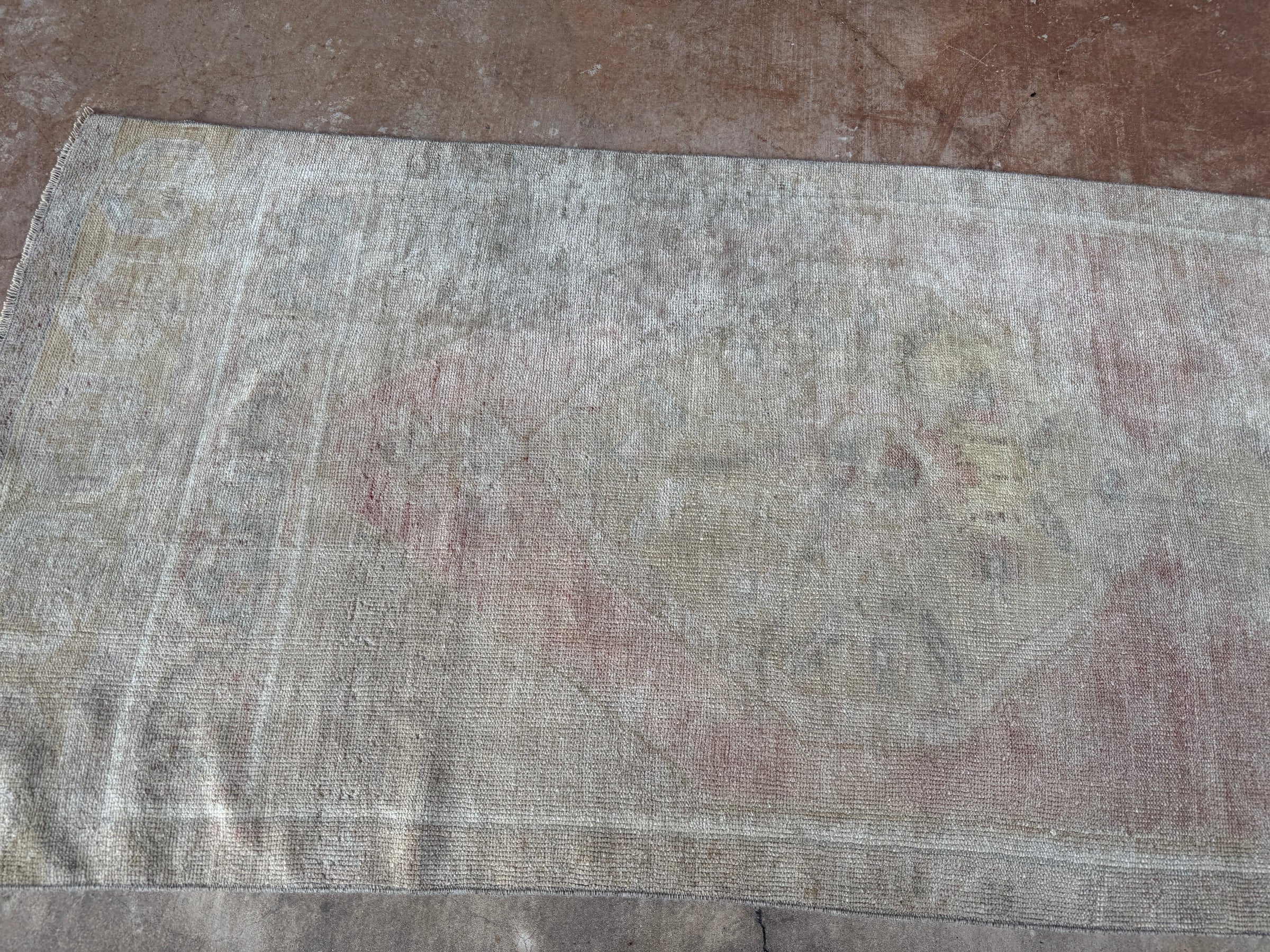 A distressed vintage-style area rug with a faded beige, gray, and muted pink pattern, laid flat on a brown textured floor.