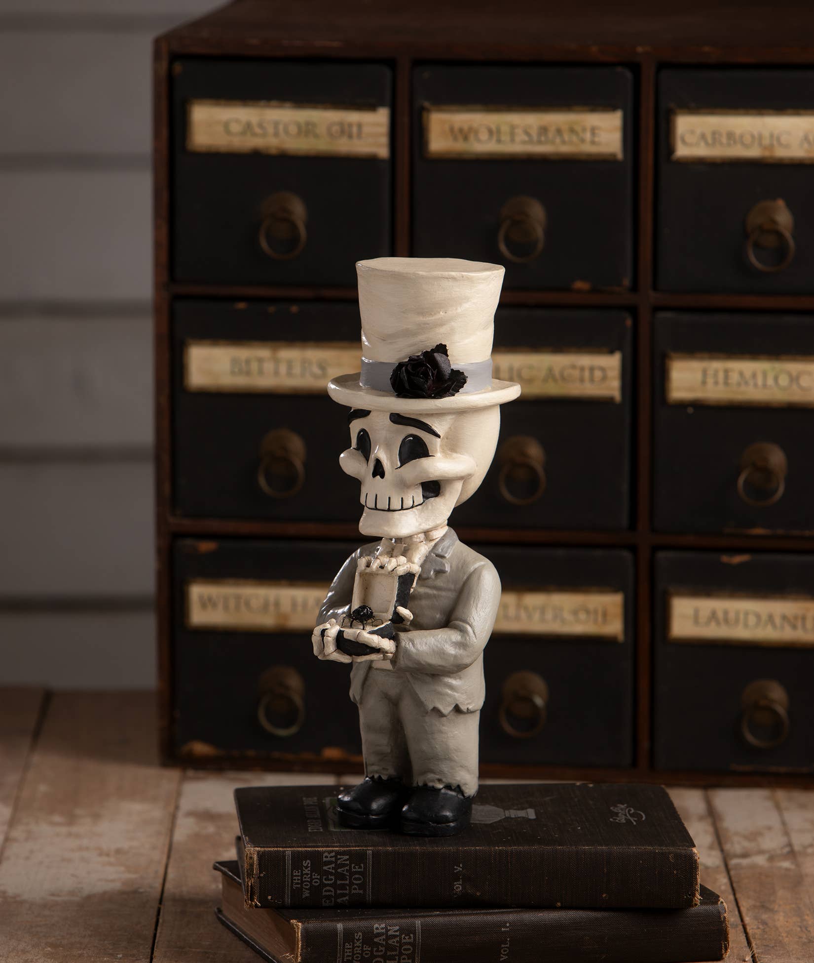 A decorative figurine of a skeleton dressed in a gray suit and white top hat with a black rose accent, standing on stacked vintage books in front of a dark wooden apothecary-style cabinet.