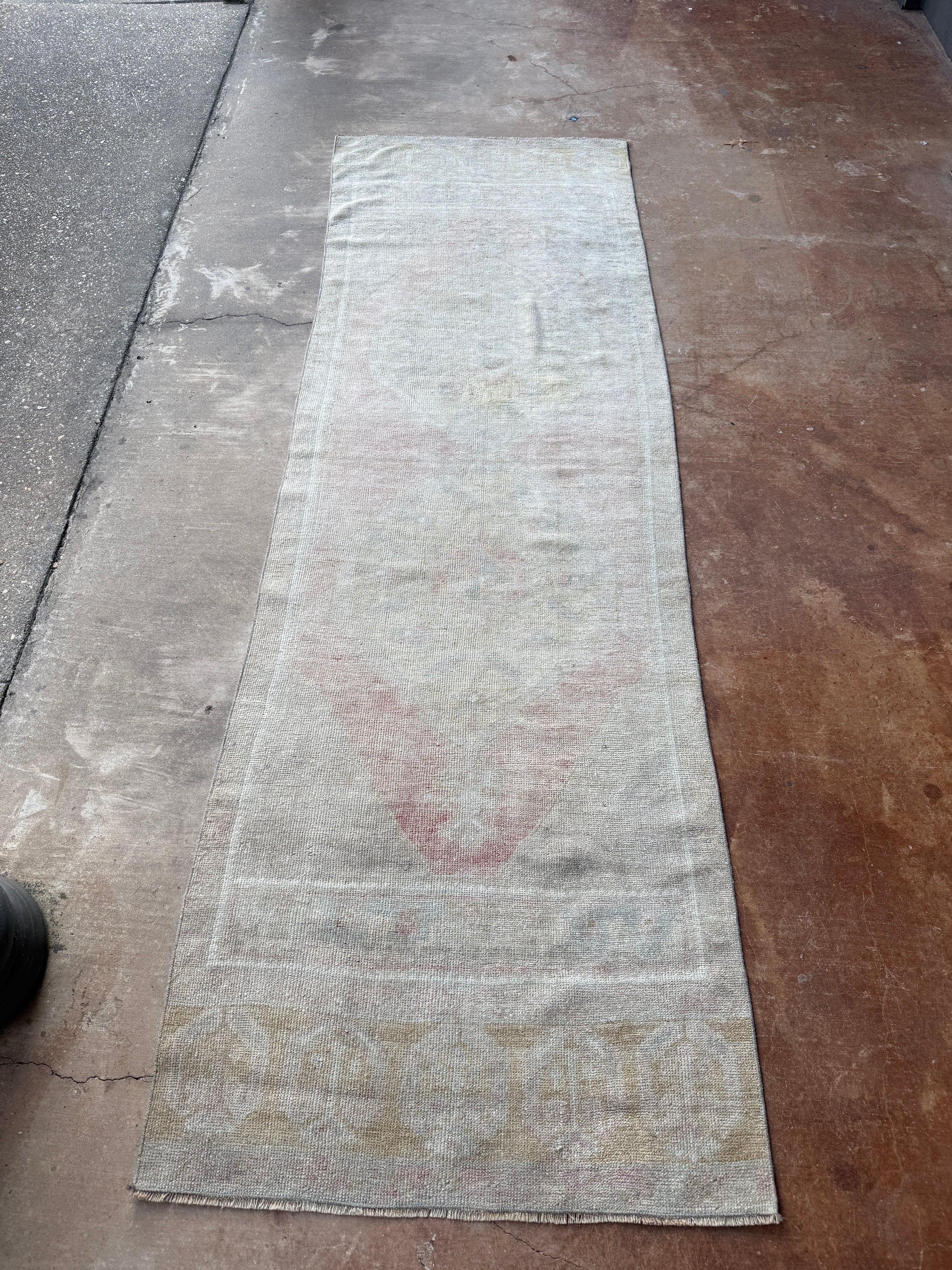 A long, narrow distressed runner rug with faded traditional patterns in muted beige, gray, and dusty red, laid flat on a concrete floor.