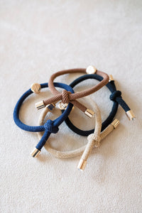 Three braided hair ties in blue, brown, and beige on a light background