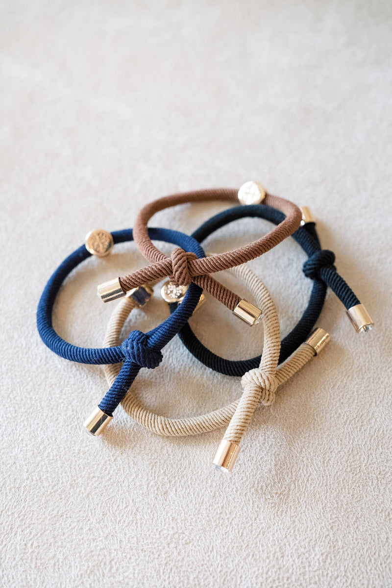 Three braided hair ties in blue, brown, and beige on a light background