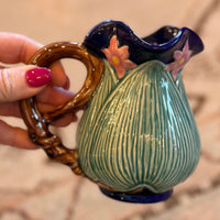 Ceramic pitcher with decorative handle held by a hand on a textured surface