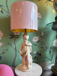Decorative lamp with a pink shade and classical figure base against a floral wallpapered wall.