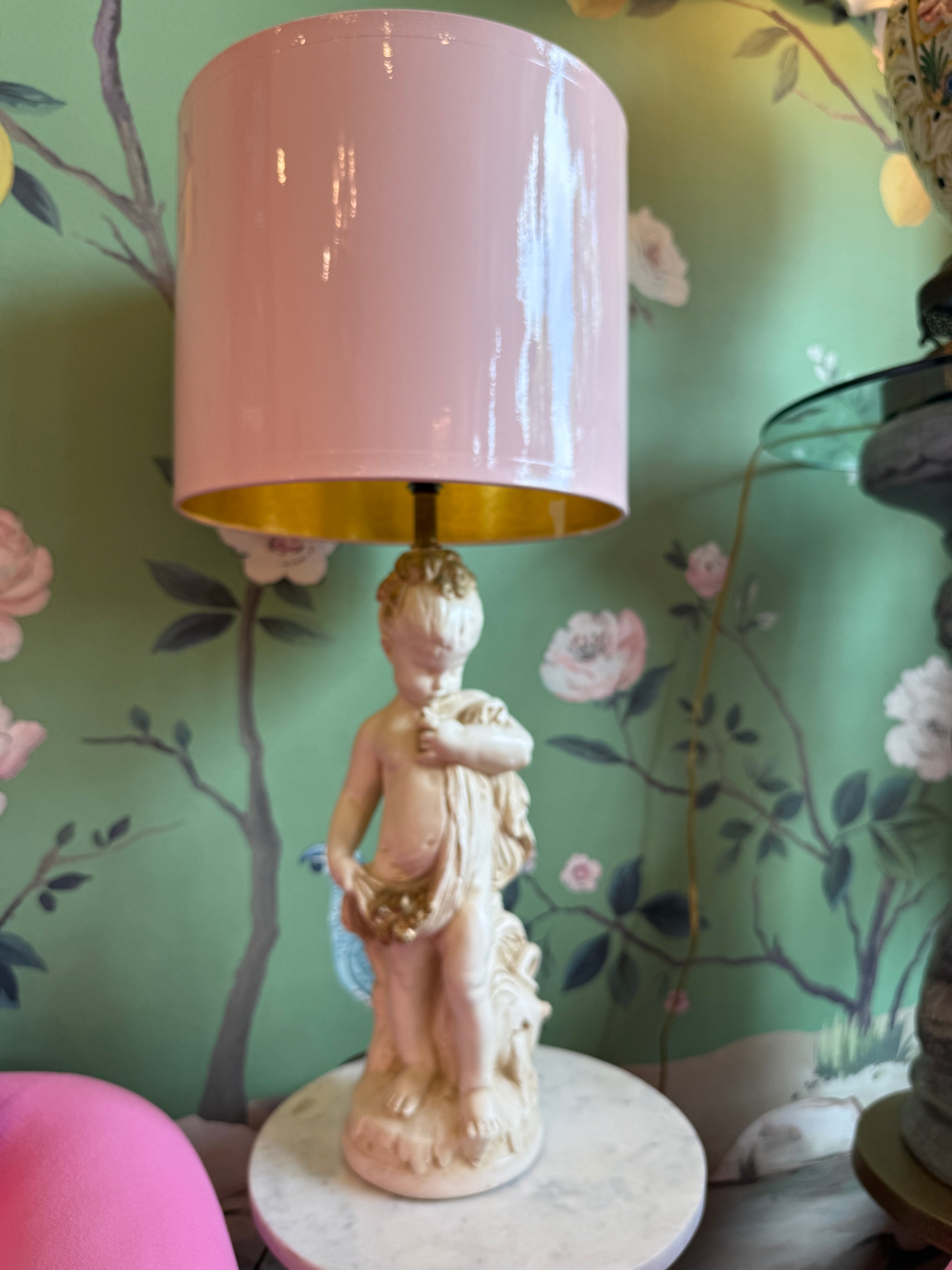 Decorative lamp with a pink shade and classical figure base against a floral wallpapered wall.