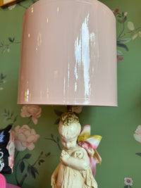 Lamp with a pink shade and decorative figure against a floral wallpapered wall.