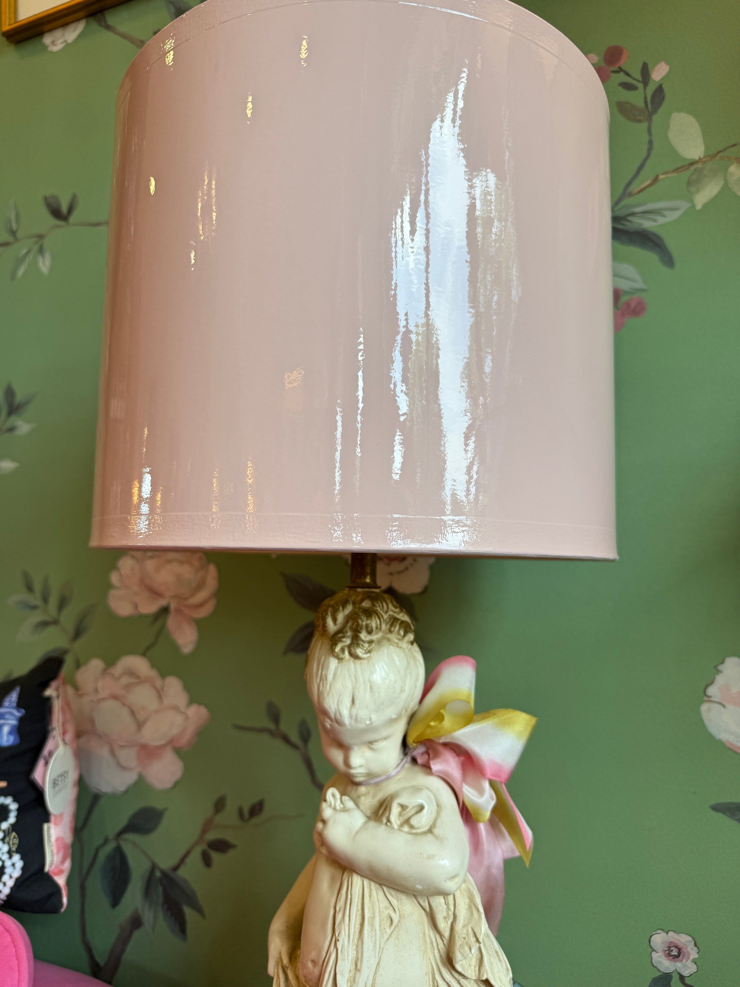 Lamp with a pink shade and decorative figure against a floral wallpapered wall.