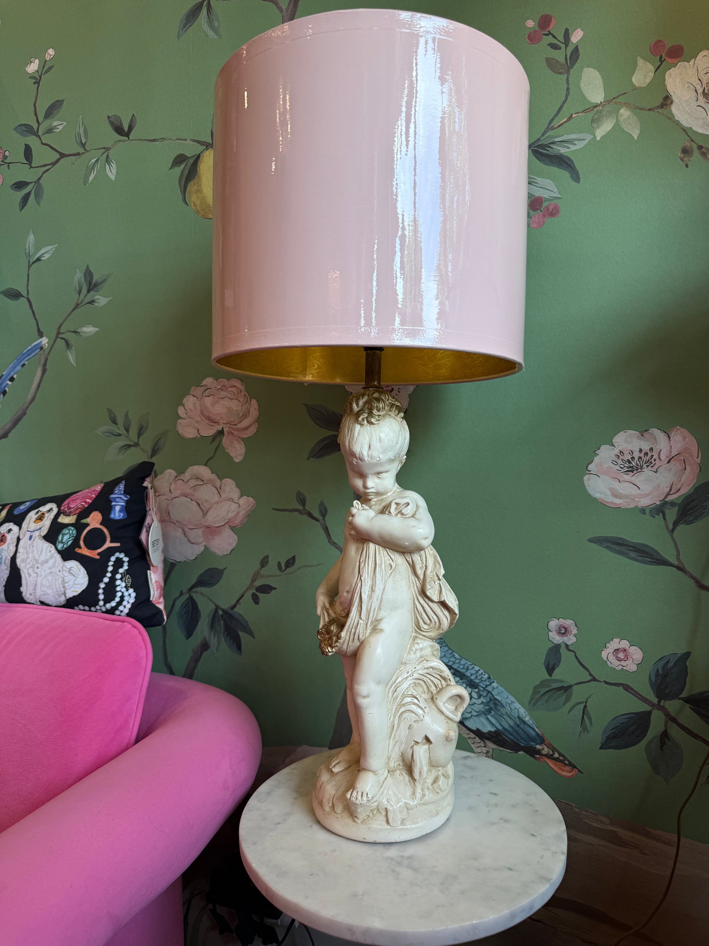 Decorative lamp with pink shade on a marble base against a floral wallpapered wall.