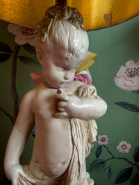 Statue of a cherub holding a heart against a floral wallpapered wall.